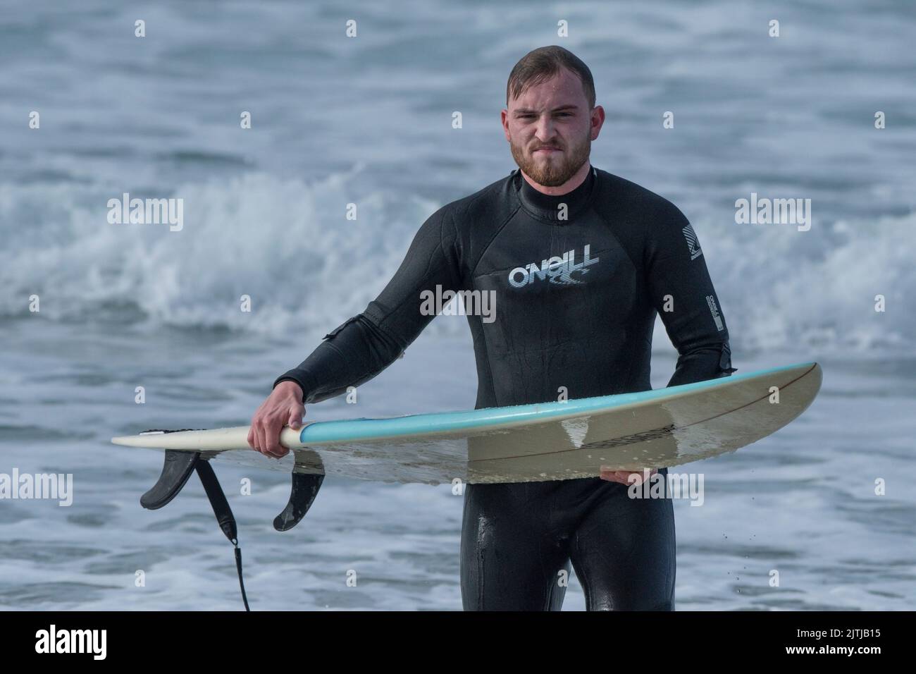 A grimacing surfer carrying his surfboard walking out of the sea at Fistral in Newquay in Cornwall in the UK. Stock Photo