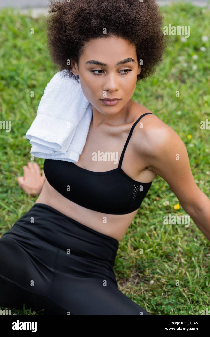 Curly African American Woman In Black Sports Bra Sitting On Lawn With White Towel On Shoulder