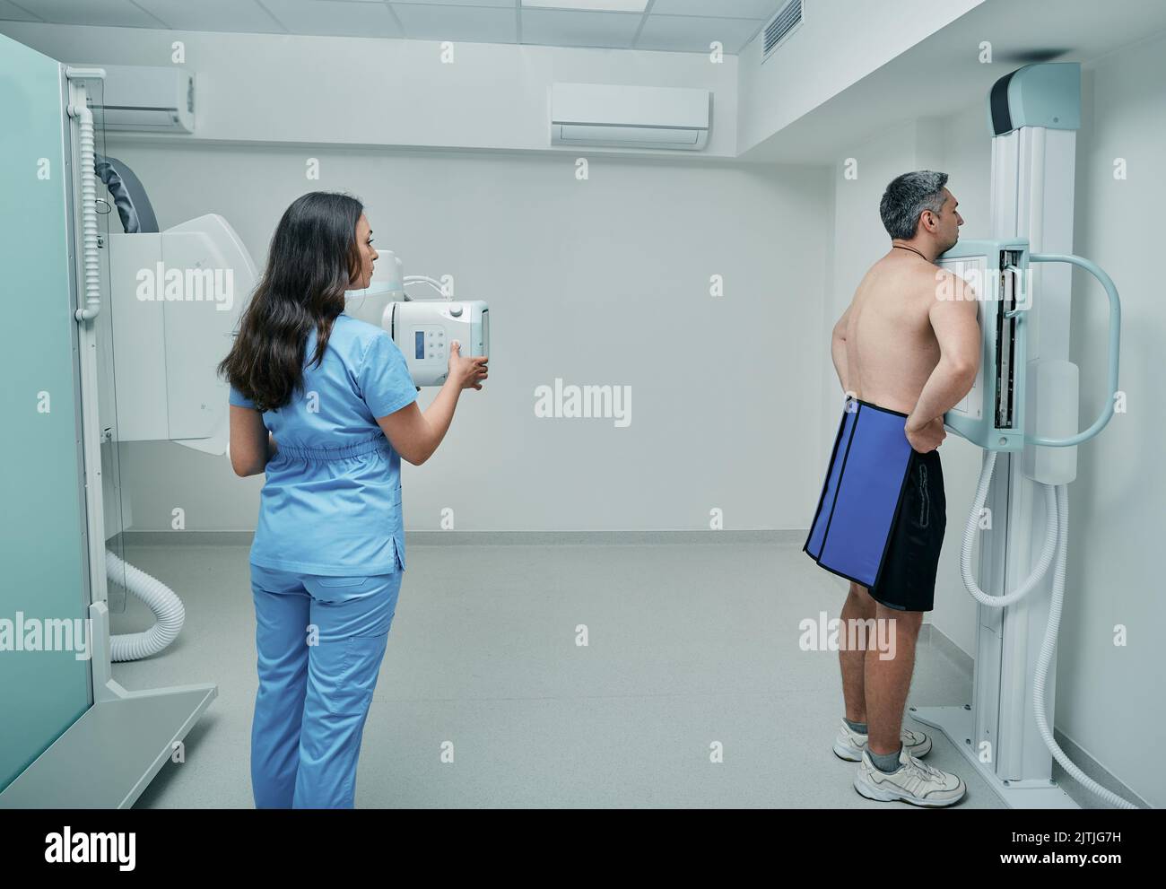 Female technician preparing male patient with protection apron for chest X-ray in hospital radiology room. Scanning to male chest, heart and lungs Stock Photo