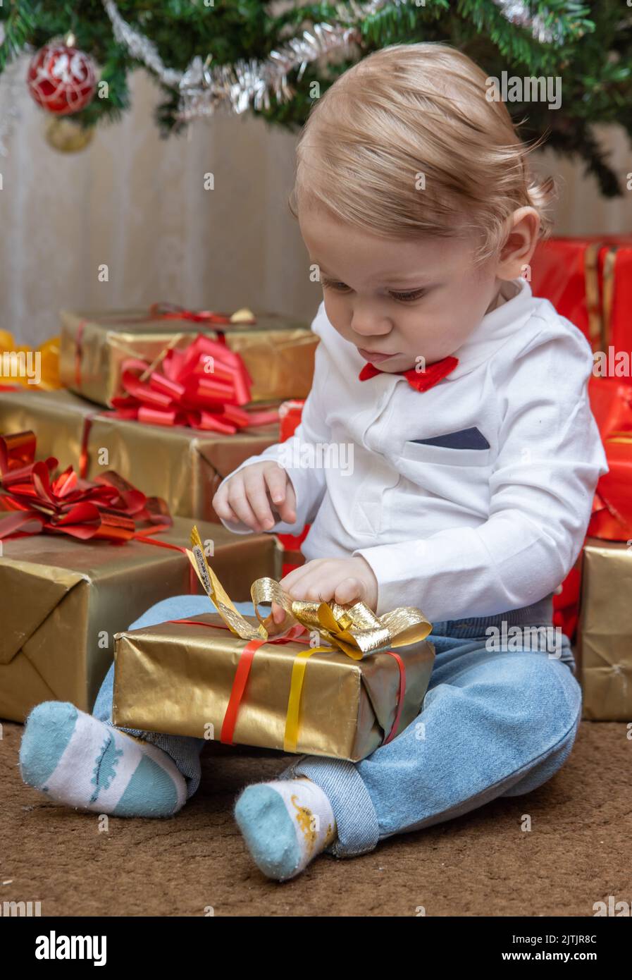 A little boy is trying to unwrap a present under the Christmas tree Stock Photo