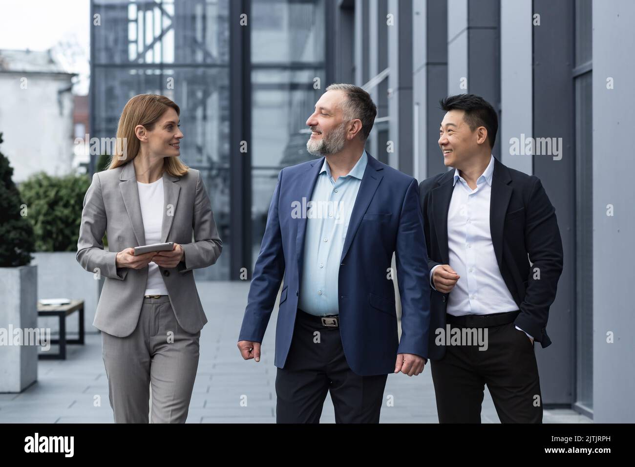 A diverse team of IT specialists, senior and experienced engineers managers team leaders, a group of three workers happily strolling outside an office building, colleagues in business suits. Stock Photo