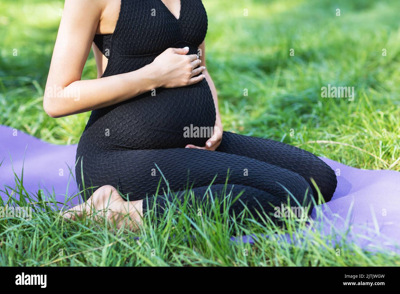 A pregnant woman in a black sports overalls is doing yoga sitting on a mat in the park on a warm summer morning Stock Photo