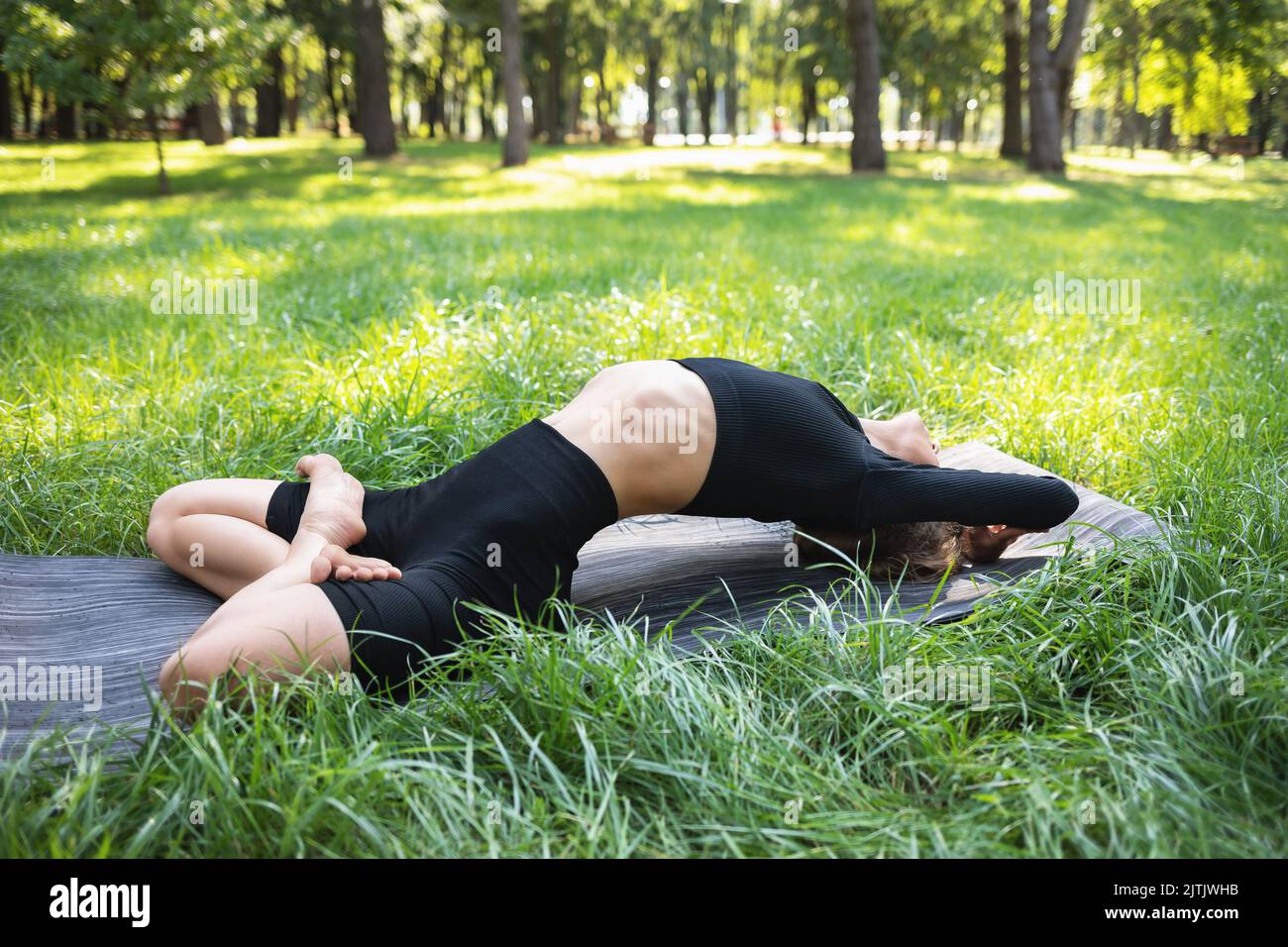 Young woman in black sportswear practicing yoga doing Matsyasana exercise, fish pose, exercising on a summer morning in the park, on a mat Stock Photo