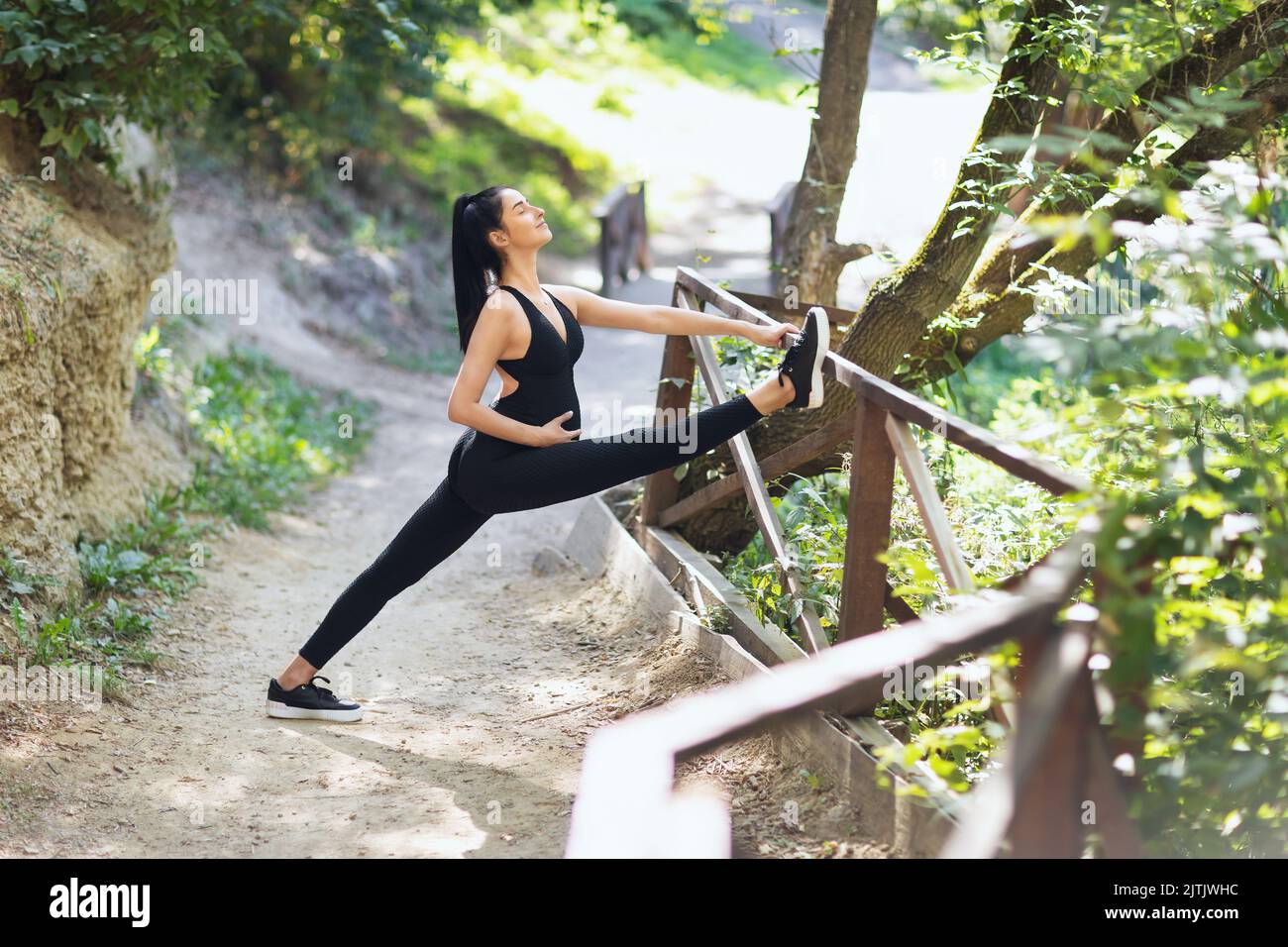 Young pregnant woman in black sports overalls practicing yoga, performs hanumanasana exercise, longitudinal twine, trains in the park on a sunny summe Stock Photo