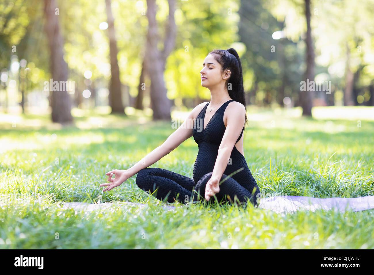 A young pregnant woman in a black sports overall practicing yoga performs meditation while sitting in a lotus position on a mat in the park Stock Photo
