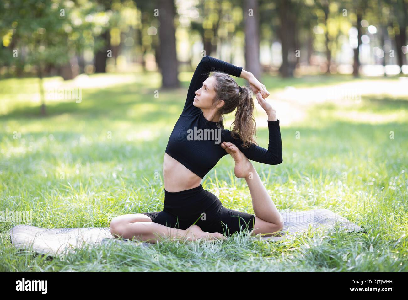Young woman in black sportswear practicing yoga doing a variation of the Eka Pada Rajakapotasana exercise, dove pose, exercising on a summer morning i Stock Photo
