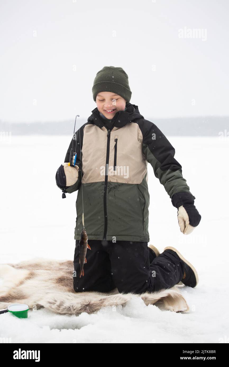 Happy boy in warm clothing kneeling while fishing on frozen lake Stock Photo