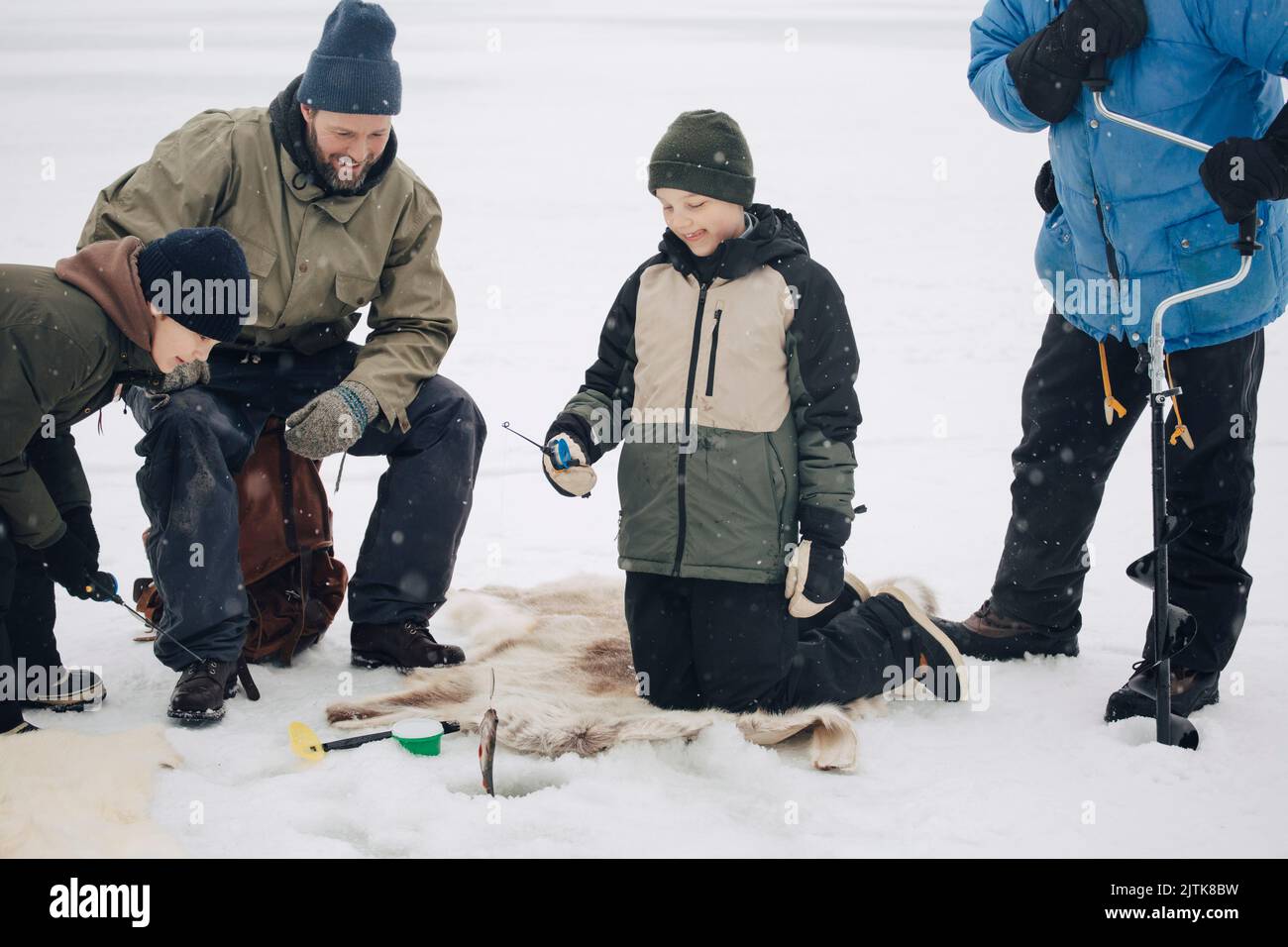 Happy boy kneeling by men and friend while fishing at frozen lake during winter Stock Photo