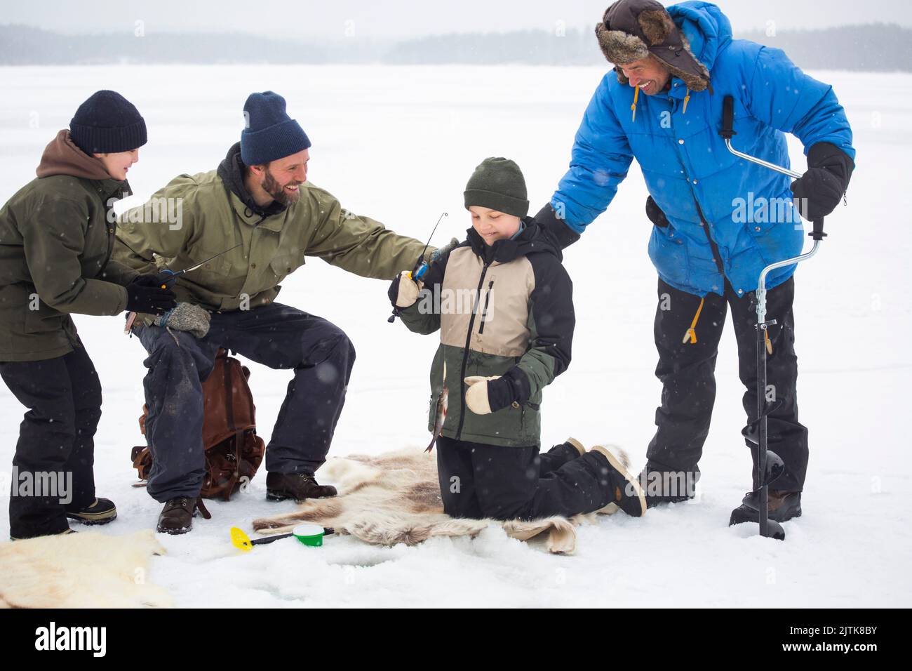 Happy mature men looking at boy fishing while kneeling on frozen lake during winter Stock Photo