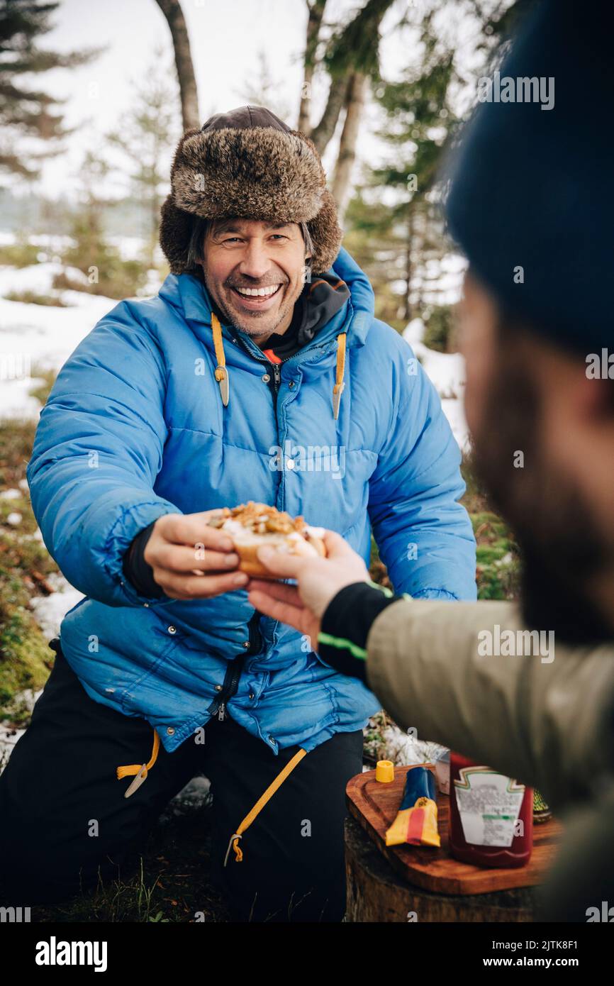 Cheerful man in warm clothing receiving hot dog from male friend Stock Photo