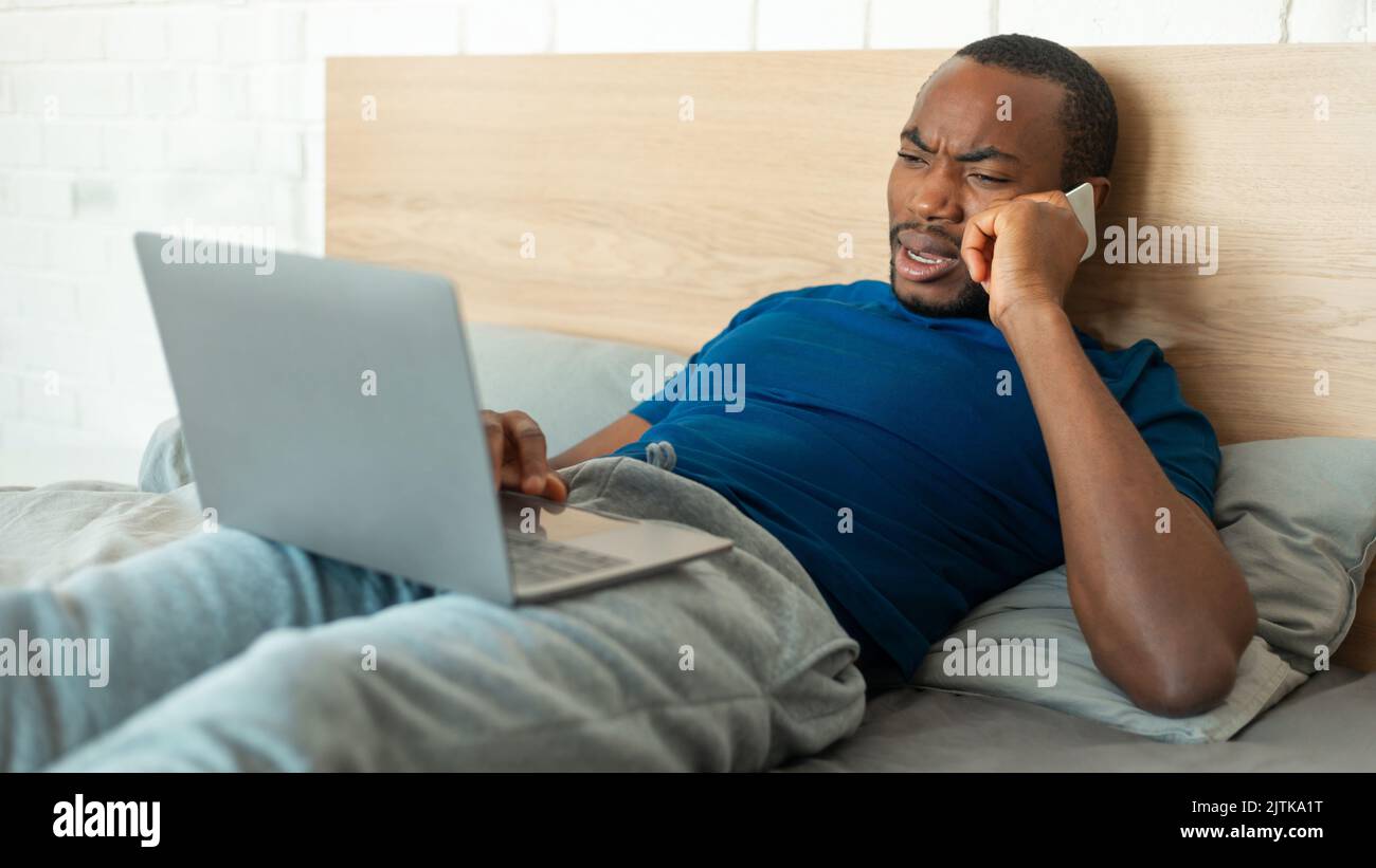 Discontented Black Guy Talking On Cellphone Using Laptop At Home Stock Photo