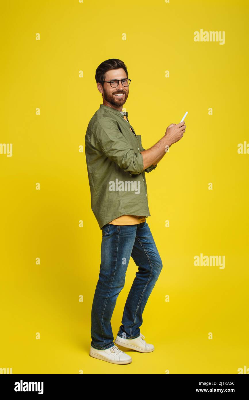 Positive caucasian man using smartphone and smiling at camera on yellow studio background, full length shot Stock Photo