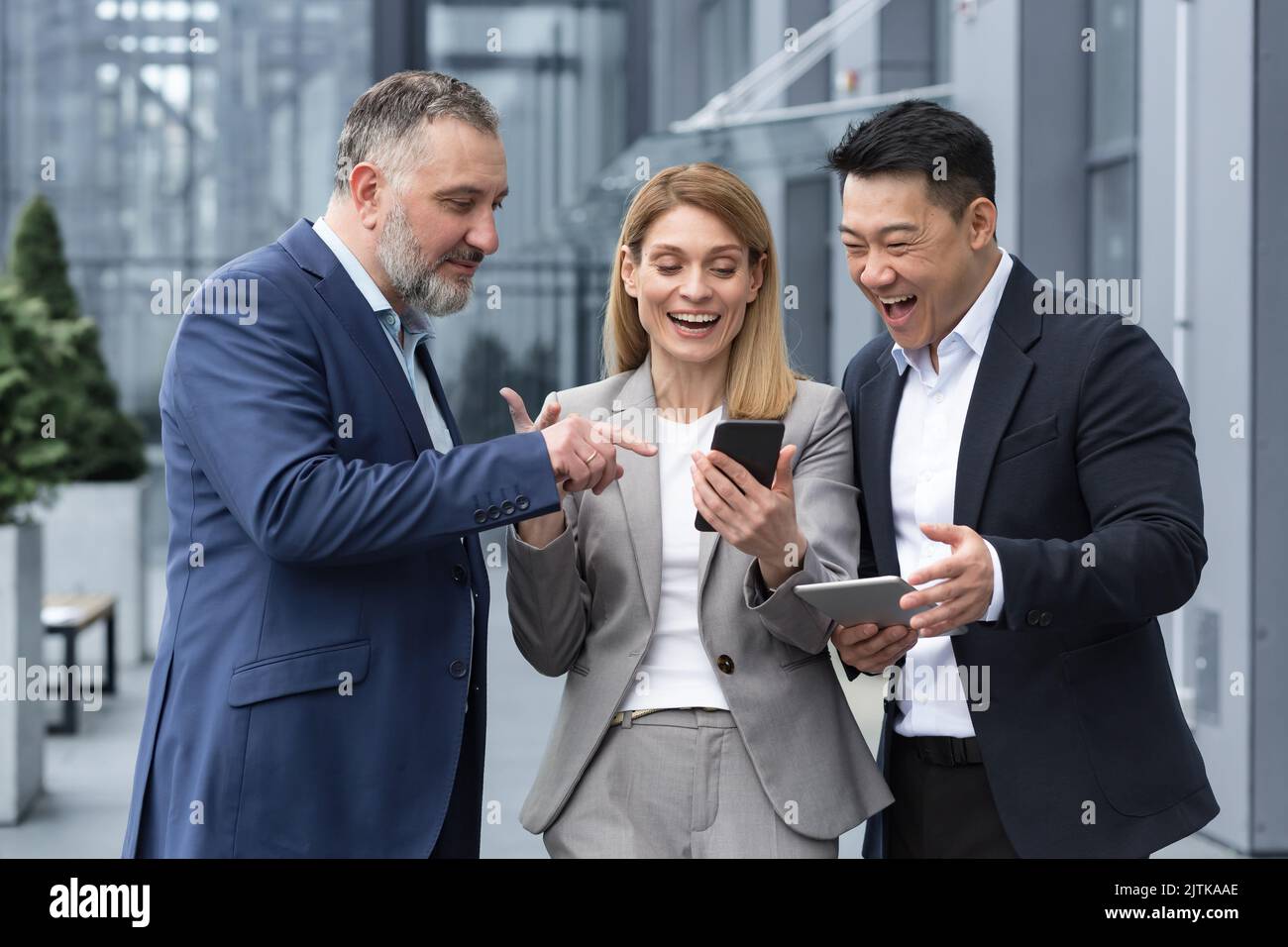 three IT specialists entrepreneurs outside office building watching video on colleague's phone, happy and smiling resting on break from work Stock Photo