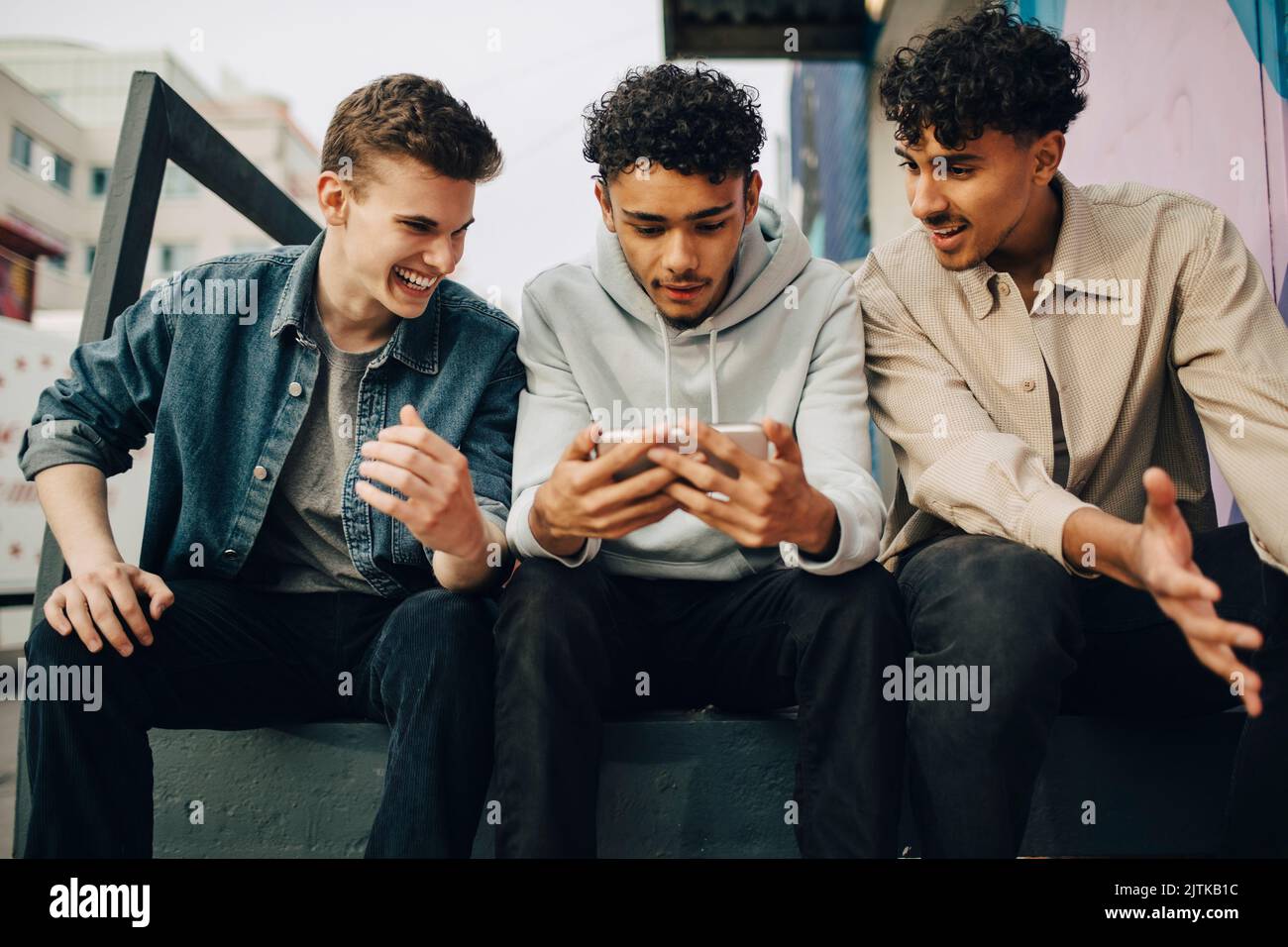 Young men looking at friend using smart phone while sitting outdoors Stock Photo