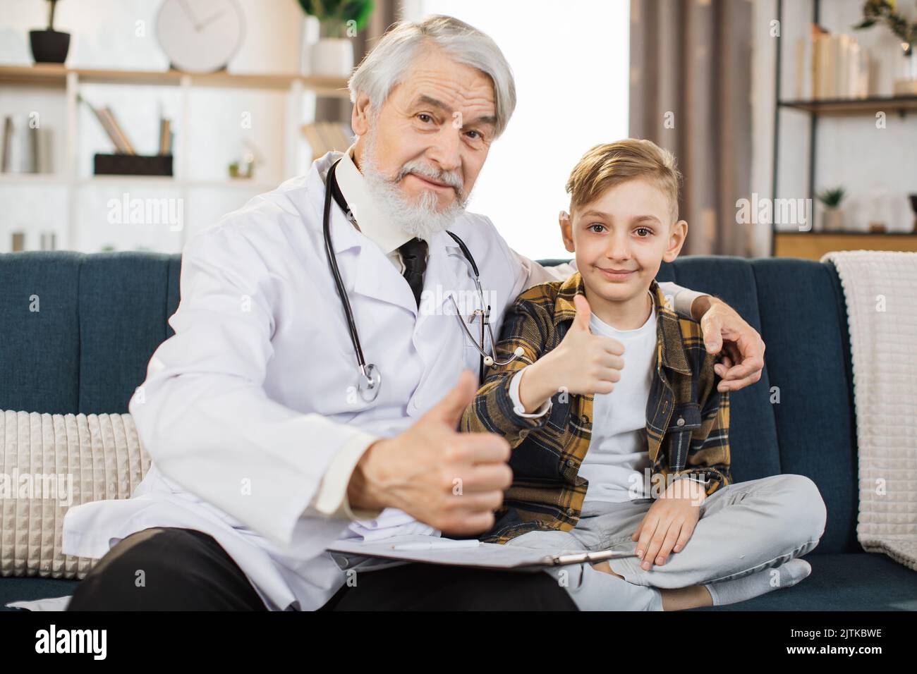 Mature man doctor in white coat and stethoscope on shoulders talking to sick boy patient, sitting on sofa, showing treatment plan, medication list, looking at camera and showing thumb up. Stock Photo