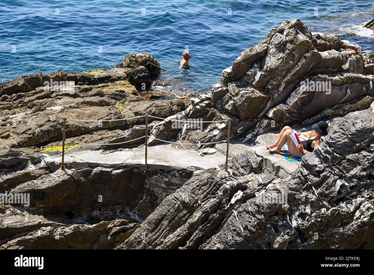 A woman reading on the cliff and a man swimming on the seashore of the Anita Garibaldi Promenade, popular tourist destination in Genoa, Nervi, Liguria Stock Photo