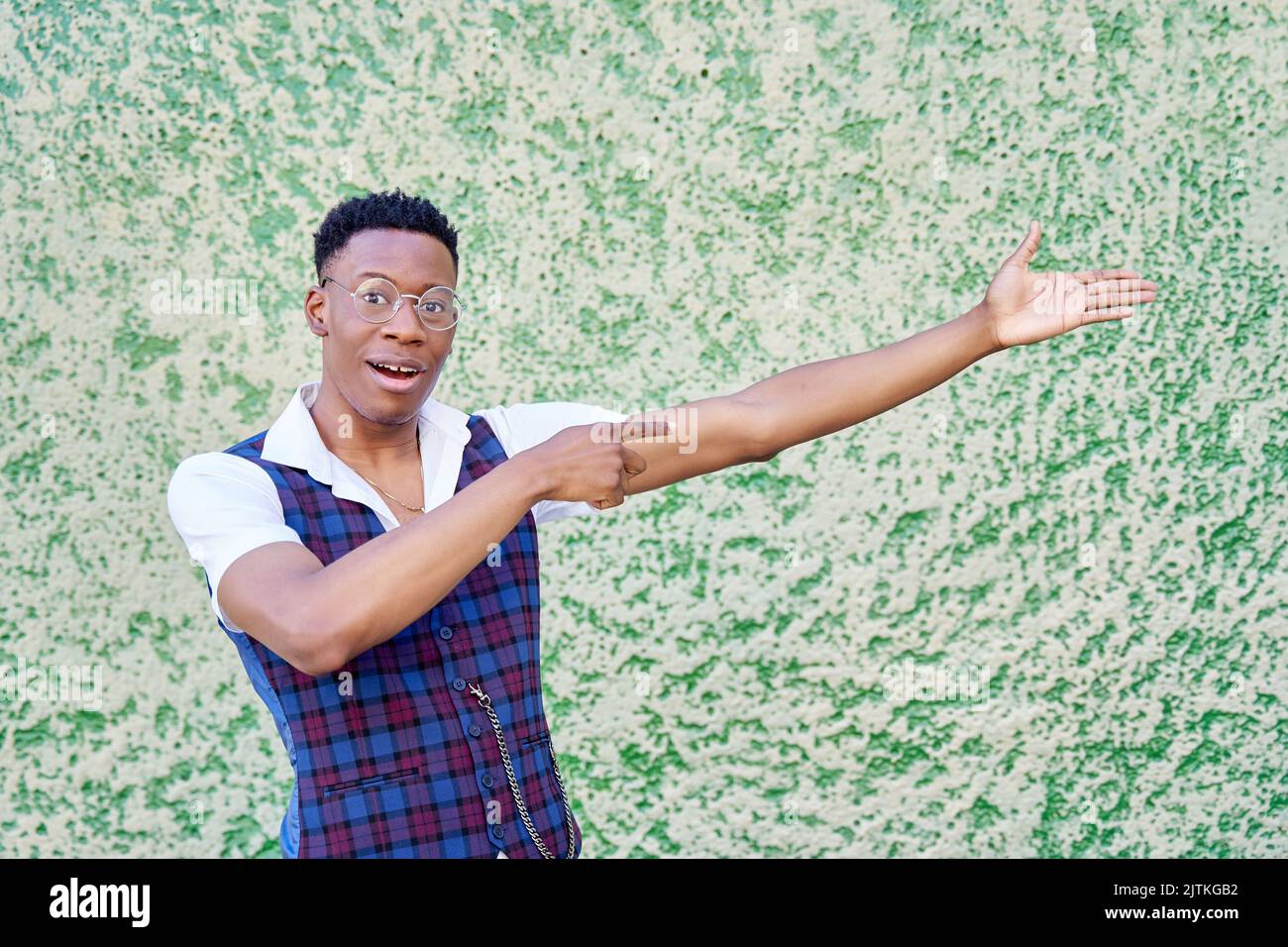 young african american man smiling cheerfully, feeling happy and pointing to the side and upwards, showing object in copy space against green wall Stock Photo