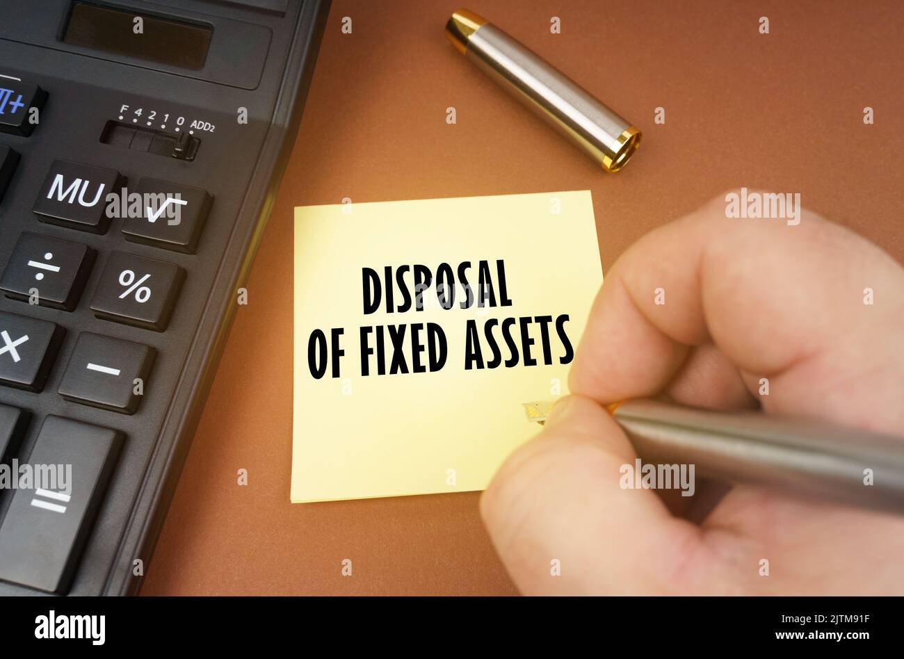 Business concept. A calculator lies on a brown surface, a hand with a pen makes an inscription on a sticker - Disposal of fixed assets Stock Photo