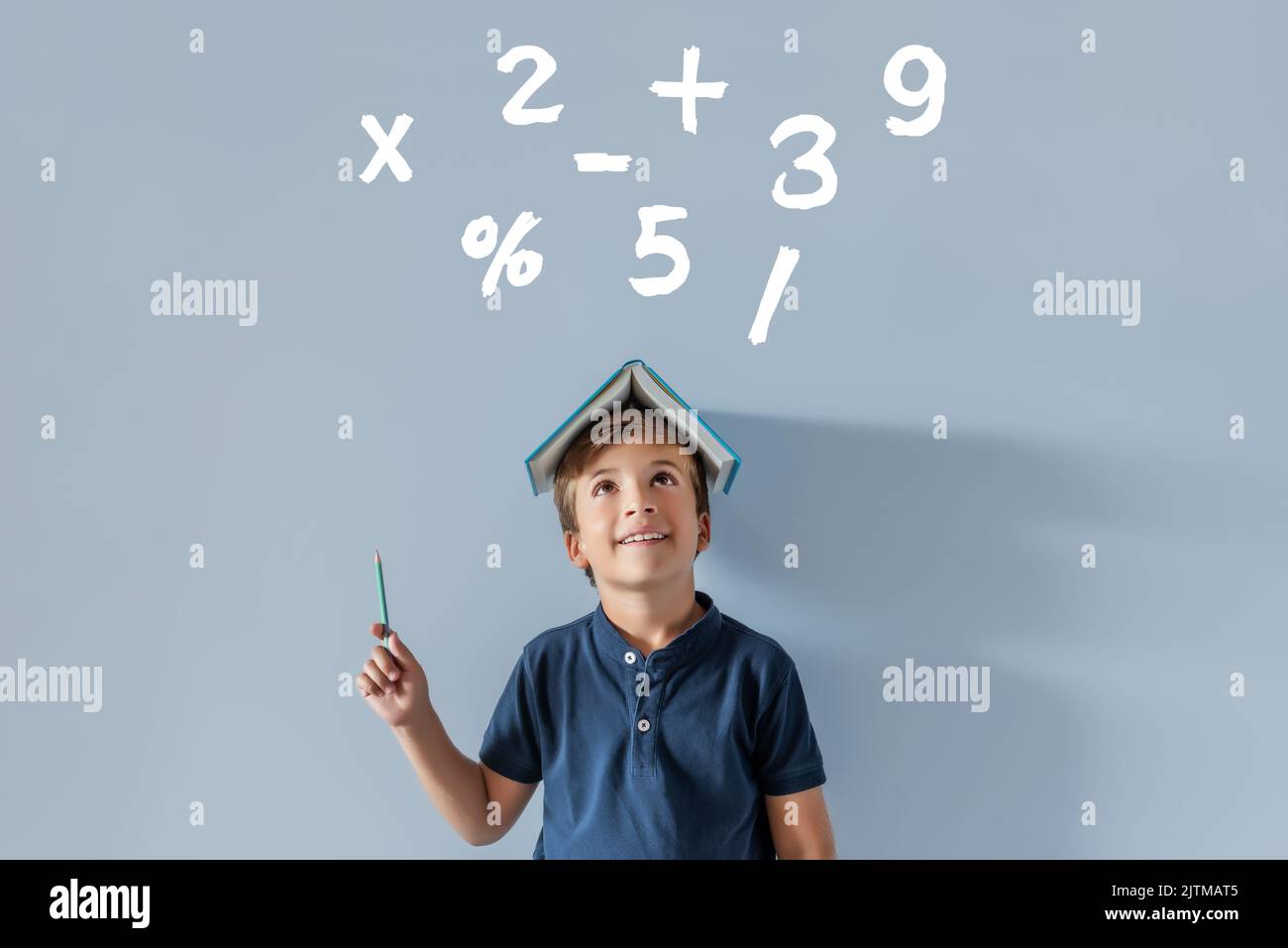 Smiling Caucasian boy with a book on his head pointing to various mathematical symbols with a pencil. Back to school, time to study subjects with numb Stock Photo