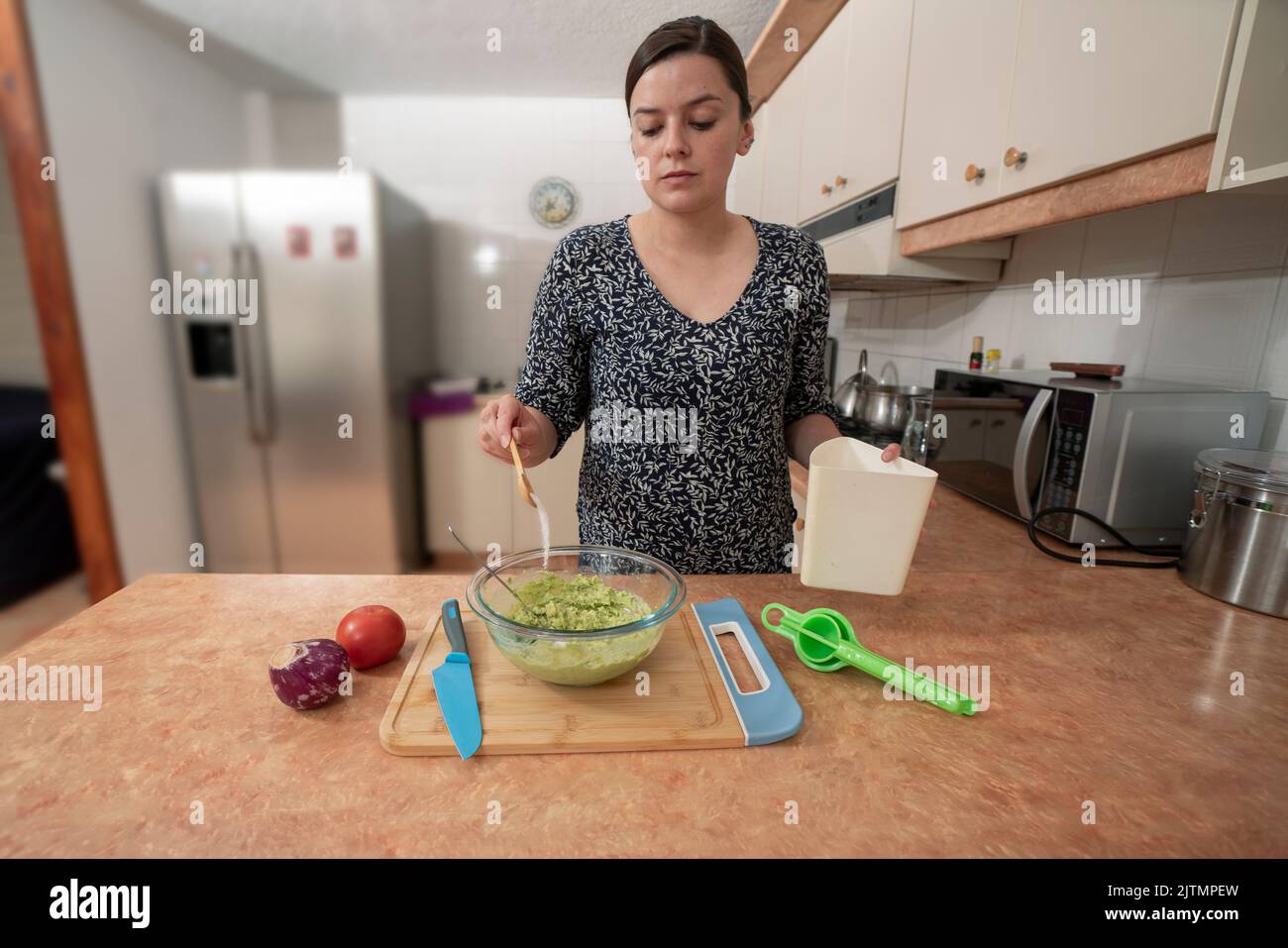 Beautiful young Hispanic woman standing with a spoon of salt seasoning the guacamole inside a glass bowl on the kitchen counter Stock Photo