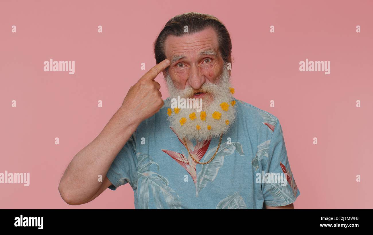 You are crazy, out of mind. Senior man with flowers in beard pointing at camera and showing stupid gesture, blaming some idiot for insane plan. Elderly grandfather alone on pink studio wall background Stock Photo