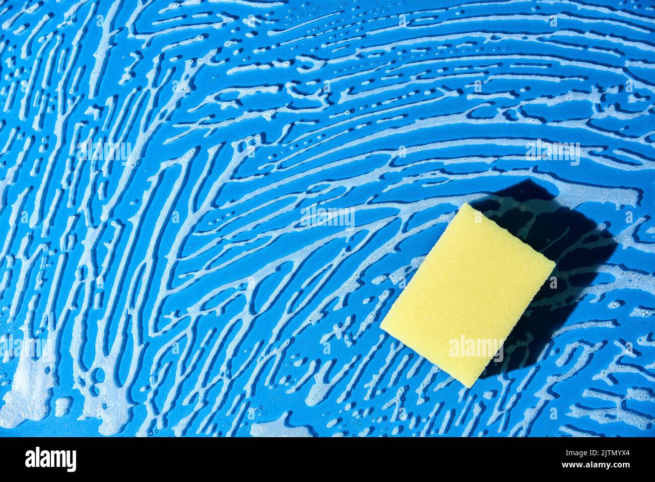 Yellow dishwashing sponge on a blue glass surface covered with soap foam. Top view composition with copy space. Concept of cleanliness. Stock Photo