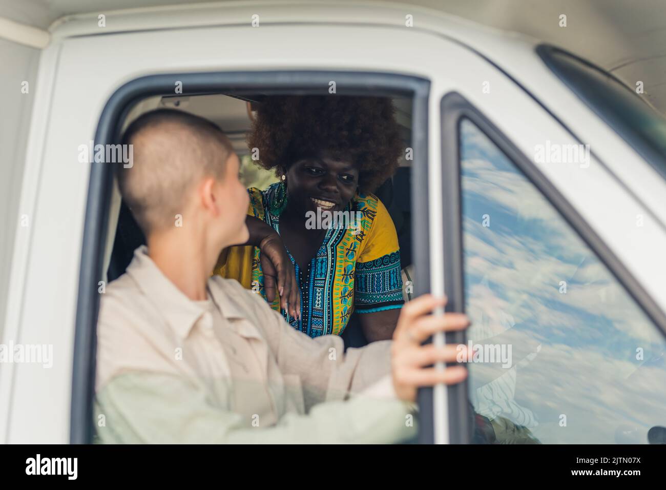 Young caucasian pretty woman with a shaved head sitting near the open window of a camping van and discussing something with her African female friend on the back seat. High quality photo Stock Photo