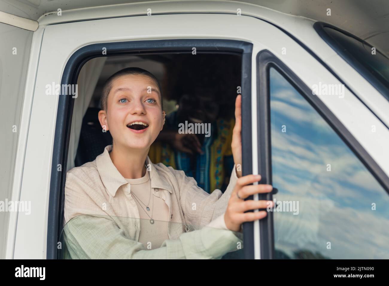 Young caucasian beautiful girl with a shaved head and her African female friend on the back seat looking out of the open window of a camping van in awe, marveling over the beautiful scenery during their road trip. High quality photo Stock Photo