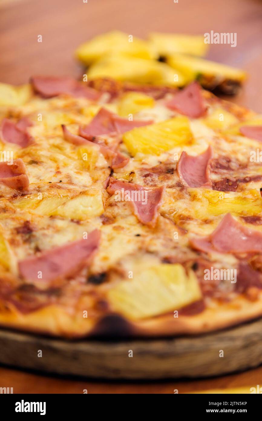 Hawaiian pizza with cheese, ham and pineapple, served on a wooden board in an Italian restaurant. Stock Photo