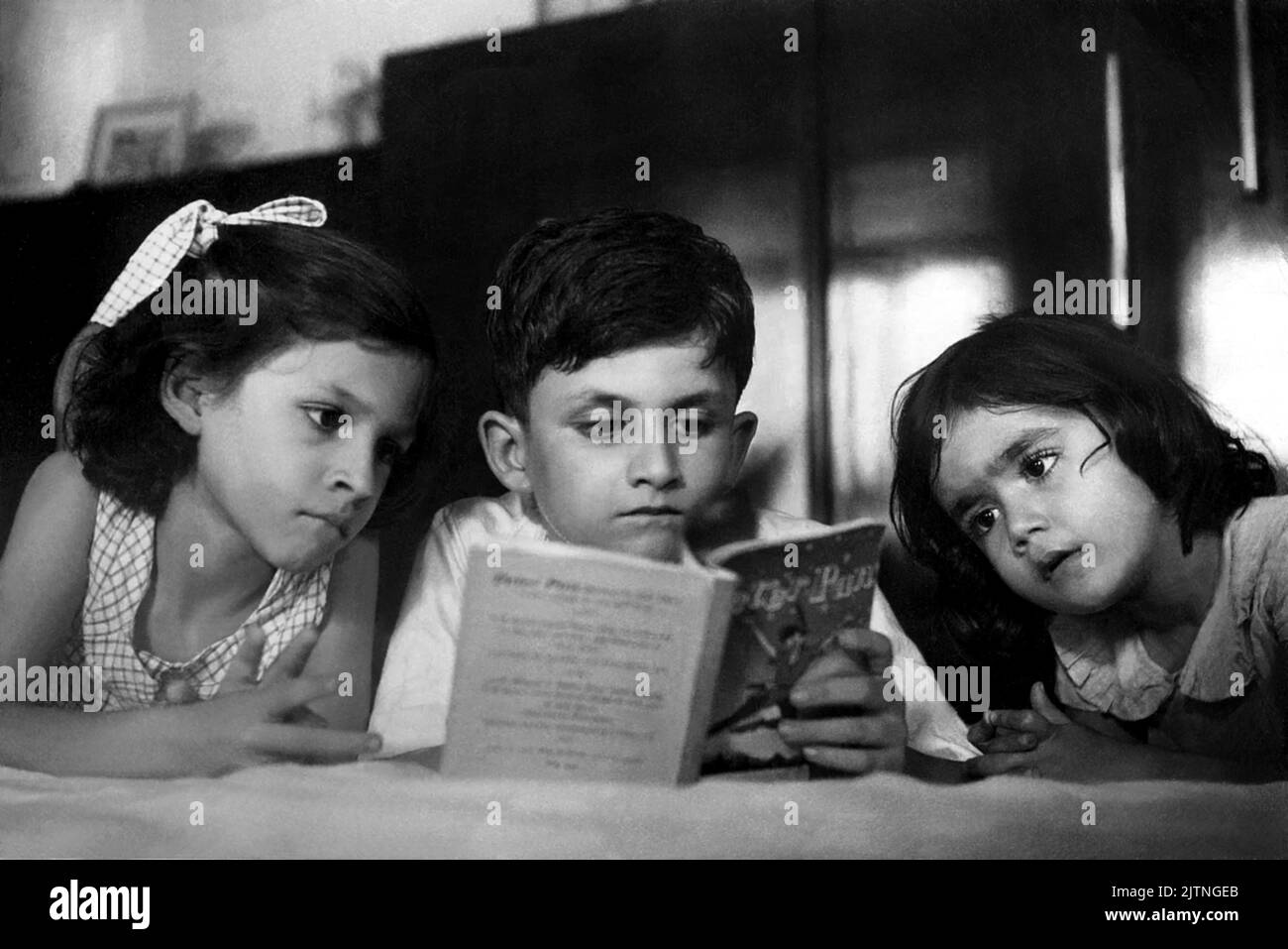 1954 , Bombay , INDIA: The celebrated indian-born british Sir SALMAN RUSHDIE ( born 19 june 1947 ) when was young boy aged 7 , reading the book PETER PAN , a fable by James Matthew Barrie , to his sisters SAMEEN and NEVID . Unknown photographer. - HISTORY - FOTO STORICHE - personalità da giovane giovani - personality personalities when was young - PORTRAIT - RITRATTO - SCRITTORE - LETTERATURA per l'INFANZIA - CHILDHOOD -CHILD - CHILDREN -  LITERATURE - lettore - libro - sorelle - sisters - brothers - fratelli - family - famiglia - FIABA - FABLE --- ARCHIVIO GBB Stock Photo