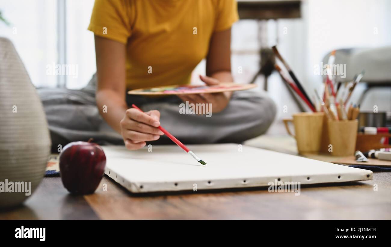 Talented female artist holding paint brush, drawing and painting her artwork on a blank canvas poster in her home living room. Hobby activity concept. Stock Photo