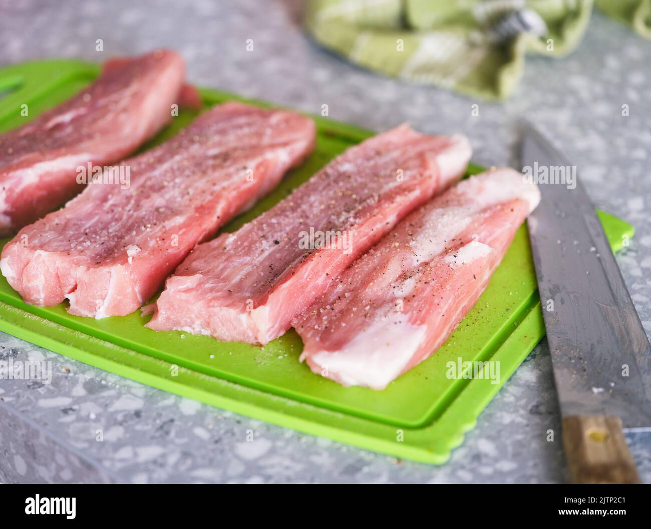 Peppered and salted raw pork slices on a green cutting board and a knife on a kitchen table Stock Photo