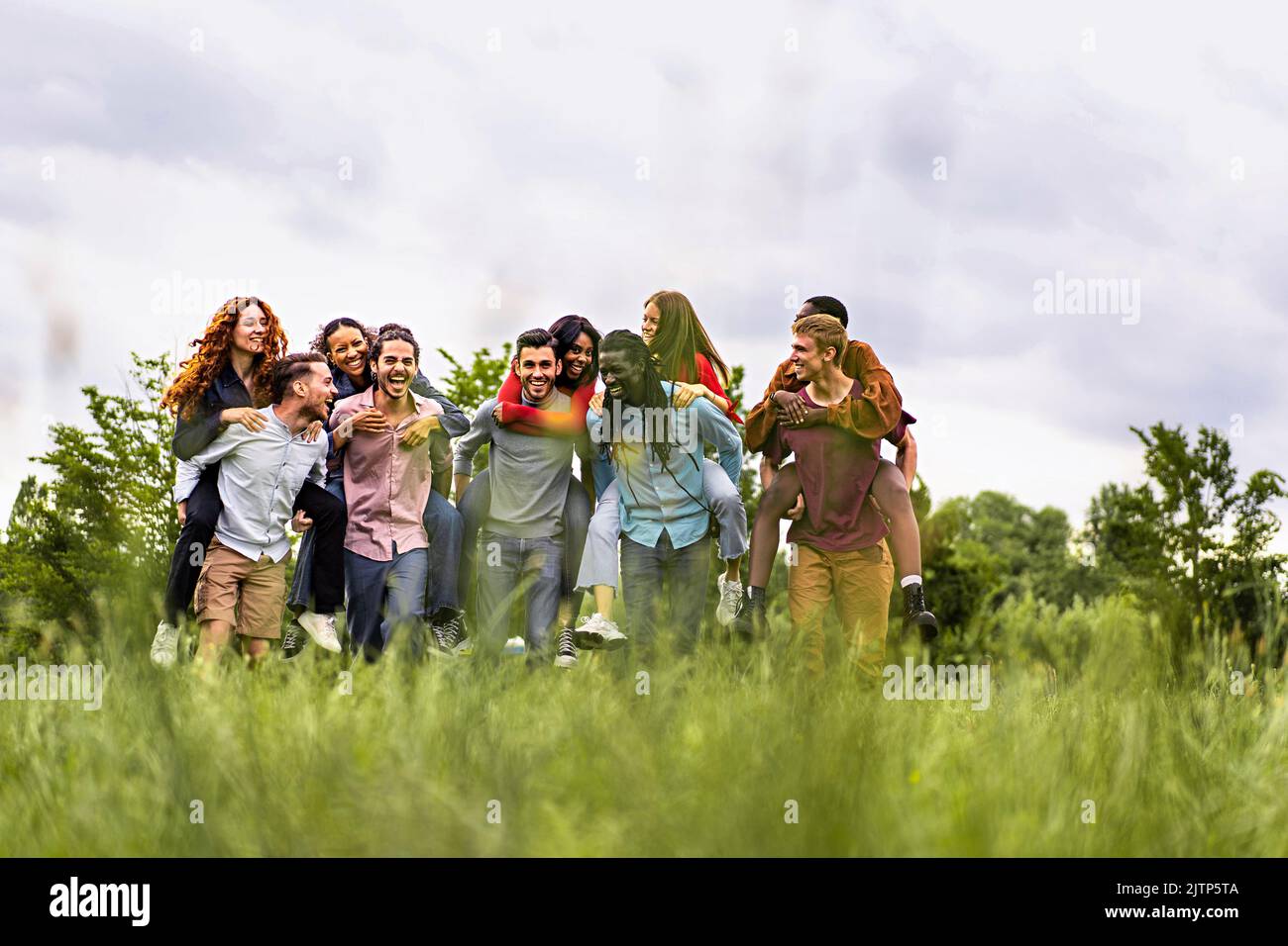 Group of multiethnic young cheerful people walking together and playing piggyback in the countryside - happy friends having fun gathering in the weeke Stock Photo
