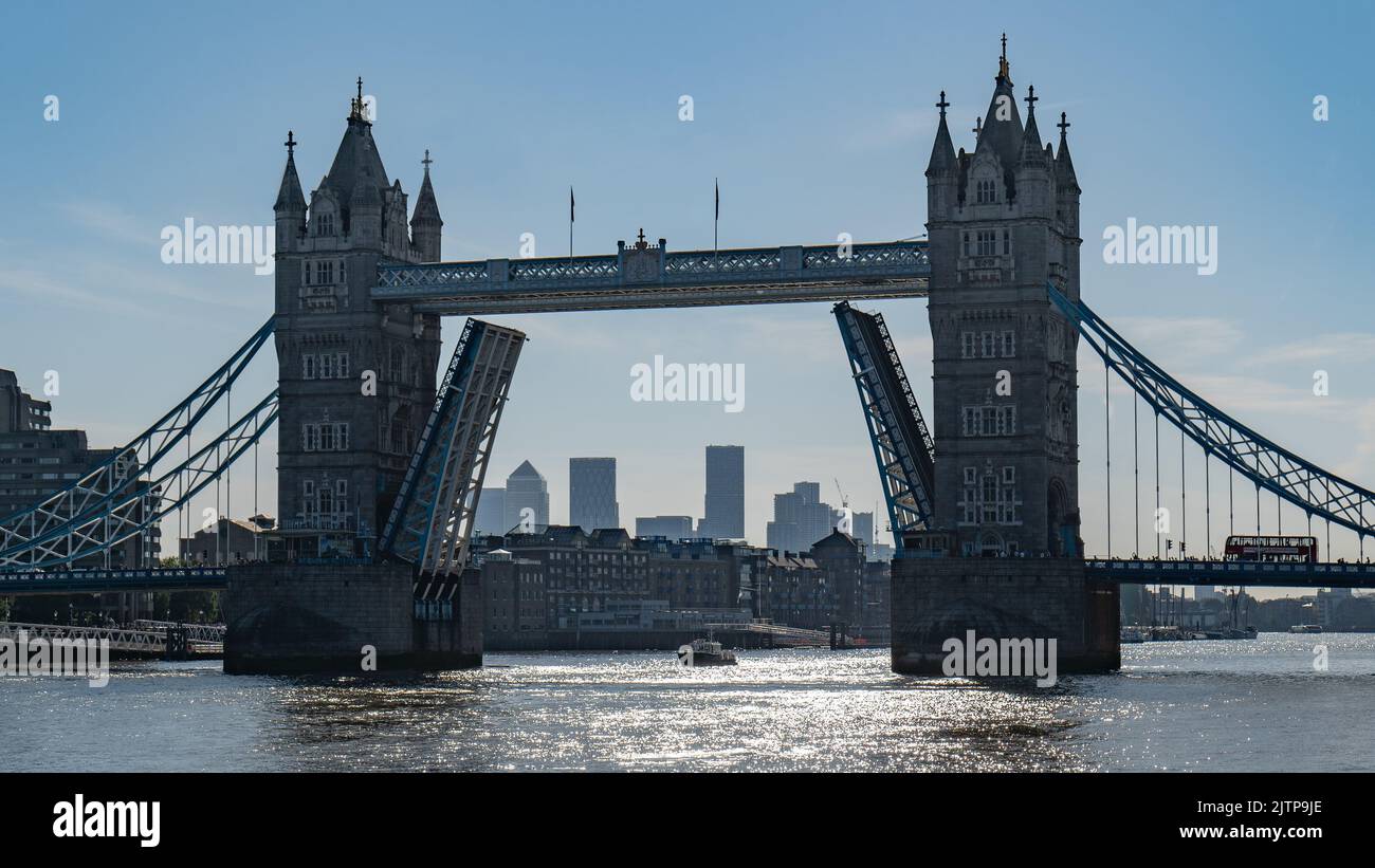An iconic raised Tower Bridge in morning sunshine with a London bus waiting to cross and Canary Wharf skyline silhouetted between the bascules. Stock Photo