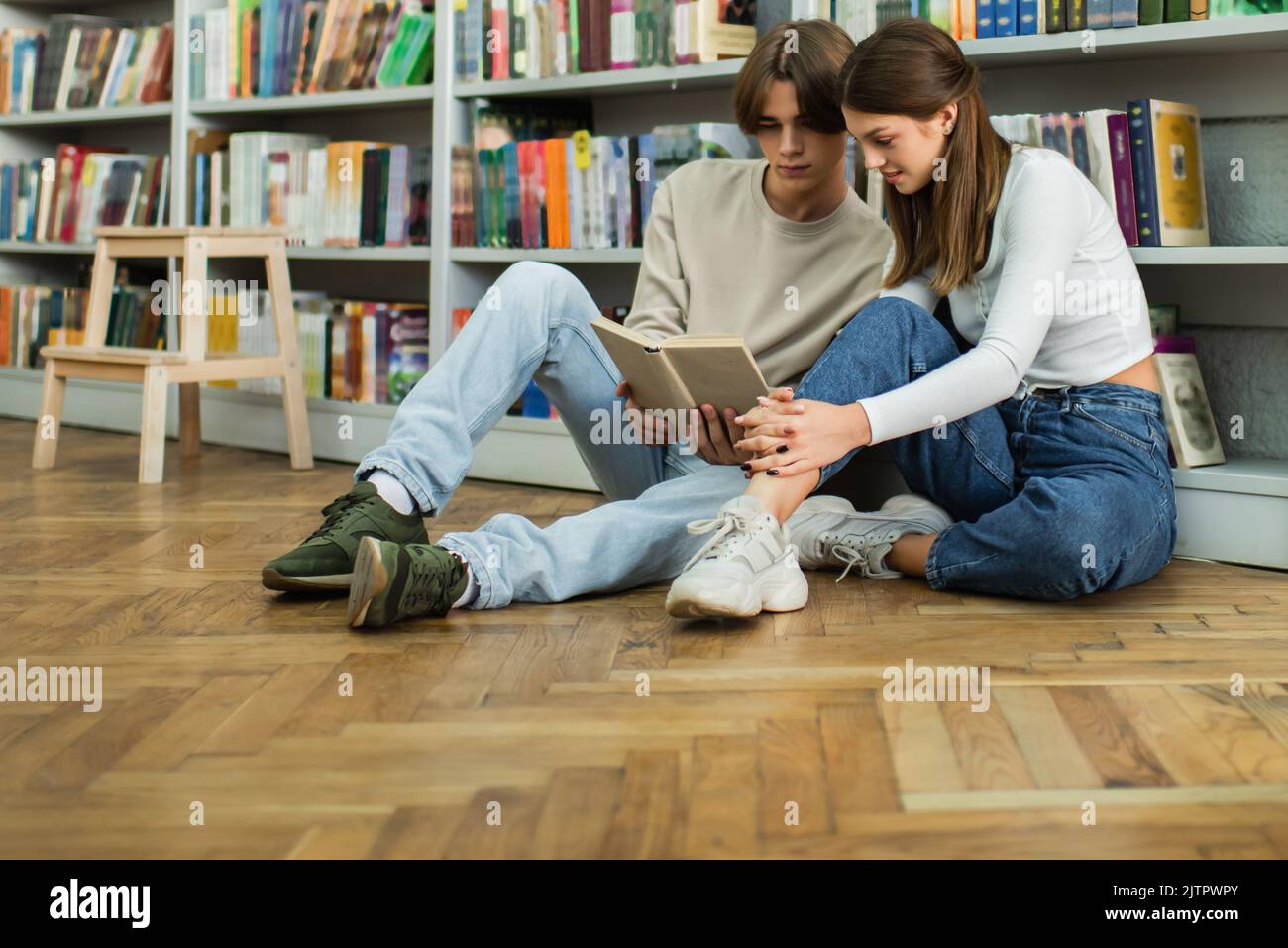 full length of teenage students sitting on floor near library racks and ...