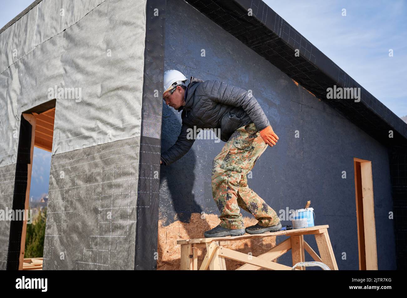 Male painter using paint roller, doing exterior paint work in a black color. Man worker building wooden frame house. Carpentry and construction concept. Stock Photo