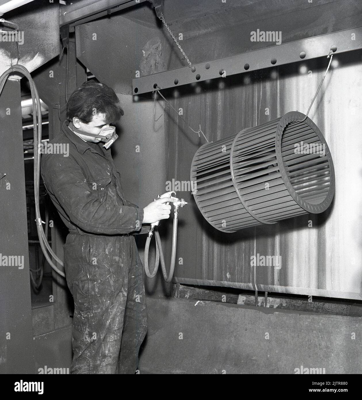 1960s, historical, a worker in overalls and nose & mouth mask using an electric spray gun to coat a metal see-thru cylinder, an industrial part, hanging in an enclosure. Stock Photo