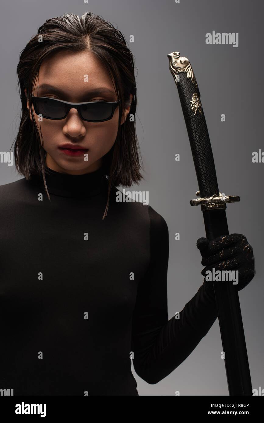 brunette asian woman in black clothes and stylish sunglasses holding katana sword in scabbard isolated on grey,stock image Stock Photo