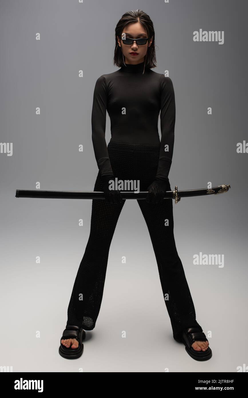 full length of asian woman in black outfit and stylish sunglasses holding katana sword on grey,stock image Stock Photo