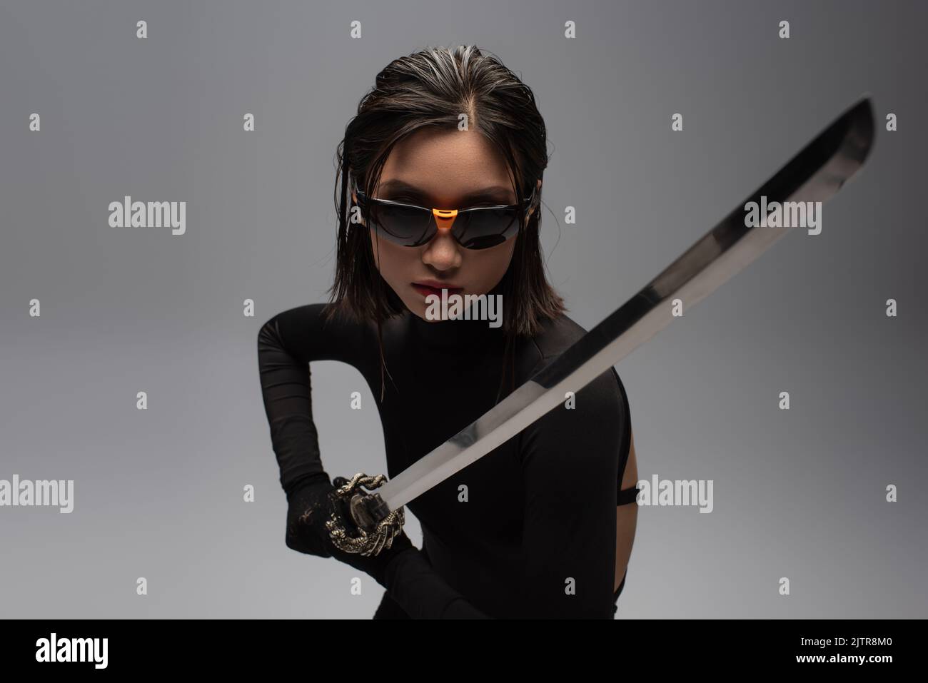 dangerous asian woman in sunglasses and black outfit holding katana isolated on grey,stock image Stock Photo