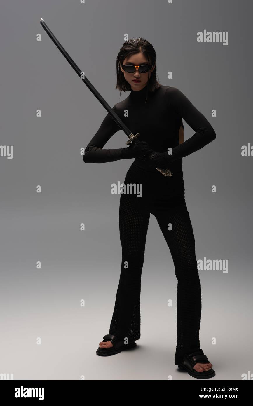 full length of young asian woman in sunglasses and black outfit holding katana on grey,stock image Stock Photo
