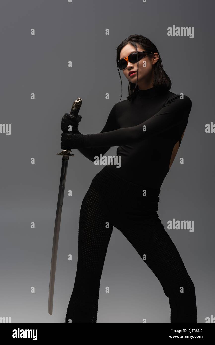 brunette asian woman in sunglasses and black clothes holding katana isolated on grey,stock image Stock Photo