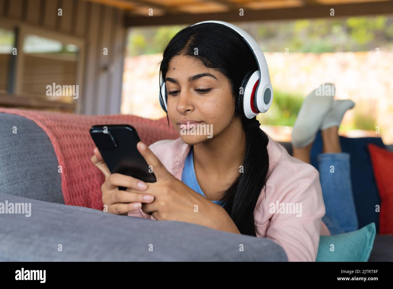 Beautiful biracial teenage girl wearing headphones and using cellphone while lying on sofa at home Stock Photo