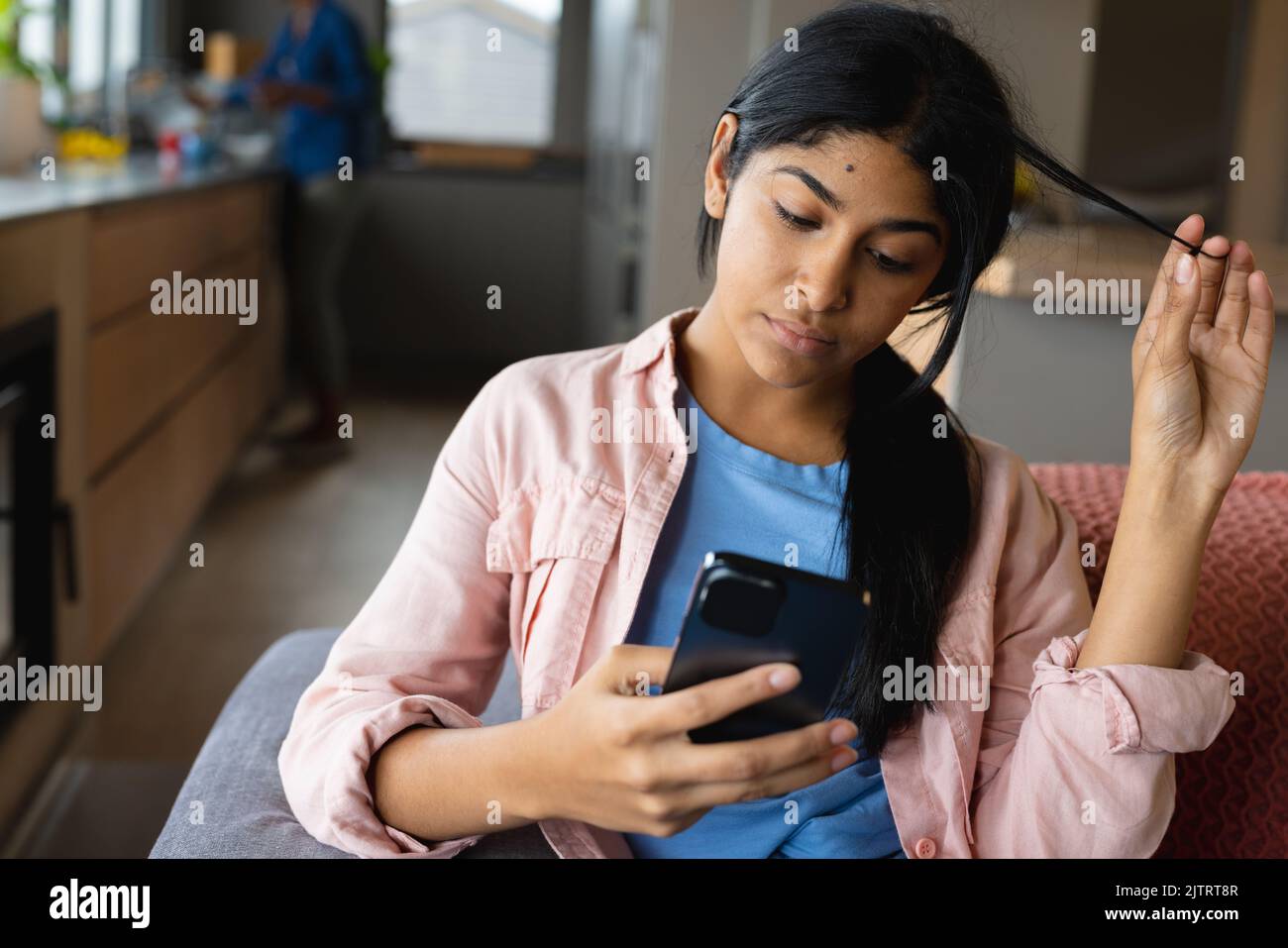 Biracial teenage girl playing with hair and using cellphone while sitting on sofa at home Stock Photo