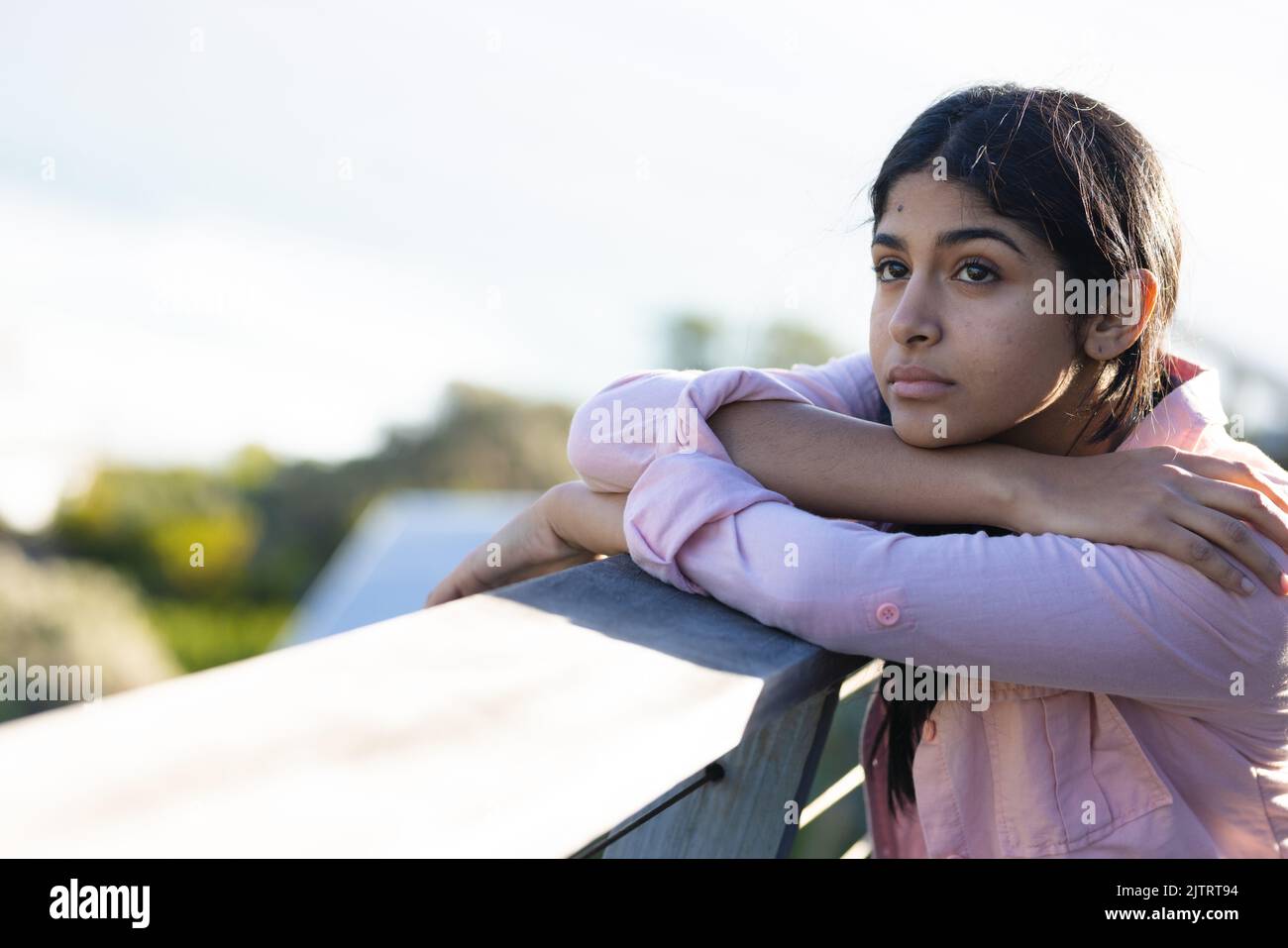 Thoughtful biracial teenage girl looking away while leaning on railing against clear sky in balcony Stock Photo