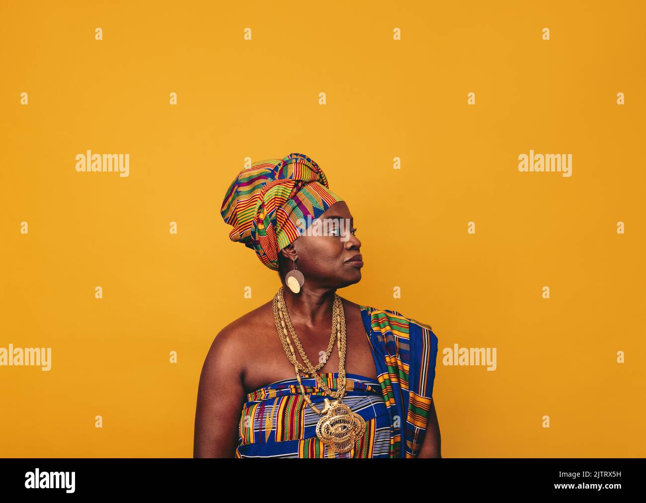 African woman wearing stylish traditional clothing against a yellow background. Mature black woman dressed in colourful Kente cloth and golden jewelle Stock Photo
