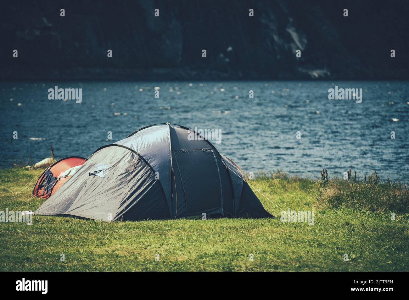 Closeup of a Tent Pitched on the Green Lake Shore with Mountain Wall in the Background. Scenic Camping Spot. Traveler Lifestyle. Outdoor Stay Theme. Stock Photo