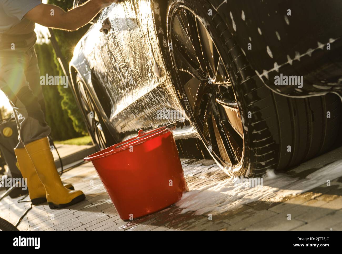 Caucasian Man in Yellow Rubber Boots Hand Washing Luxury Car on Sunny Summer Day. Red Bucket with Washing Liquid Standing Next to Rear Wheel. Vehicle Stock Photo