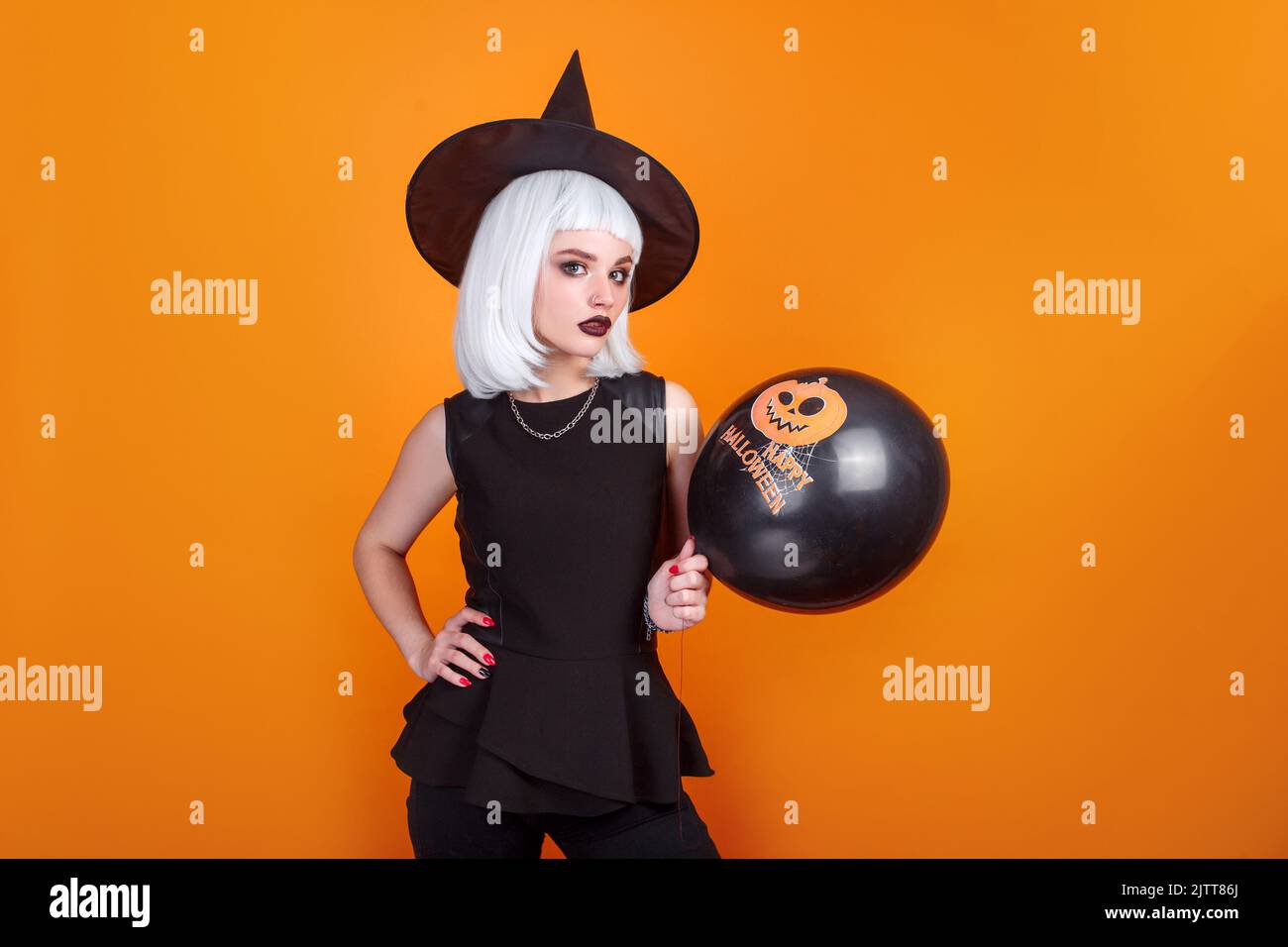 Portrait of beautiful young woman in black witch halloween costume with black balloon over orange background Stock Photo