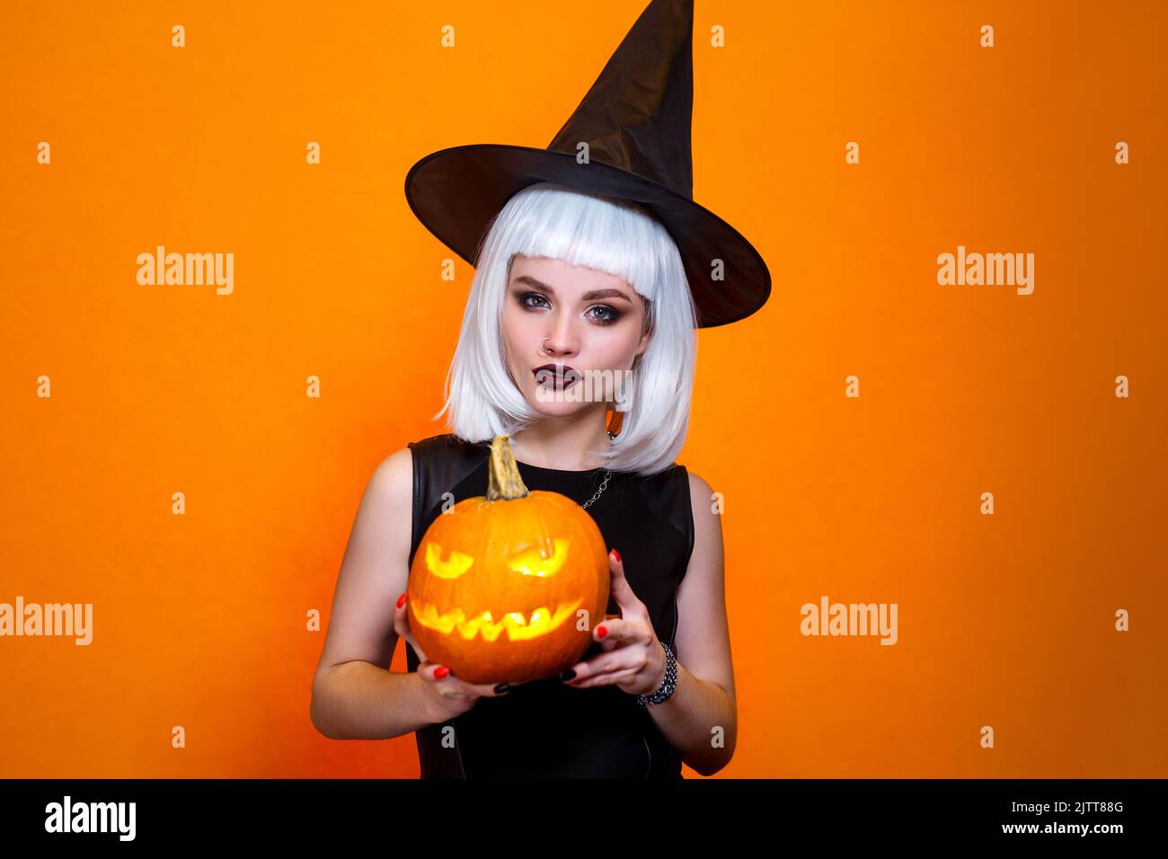 Halloween witch in hat and costume holding carved pumpkin on orange background. Stock Photo
